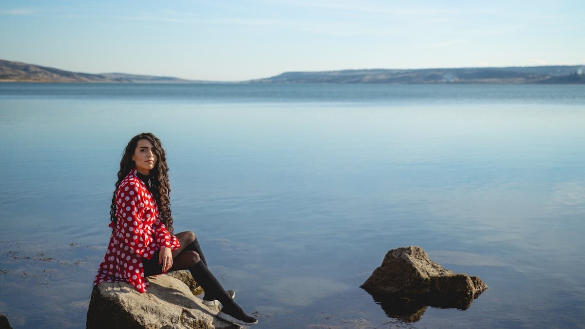 A woman in a red polka dot dress seated on rocks by a calm lake under a clear sky.